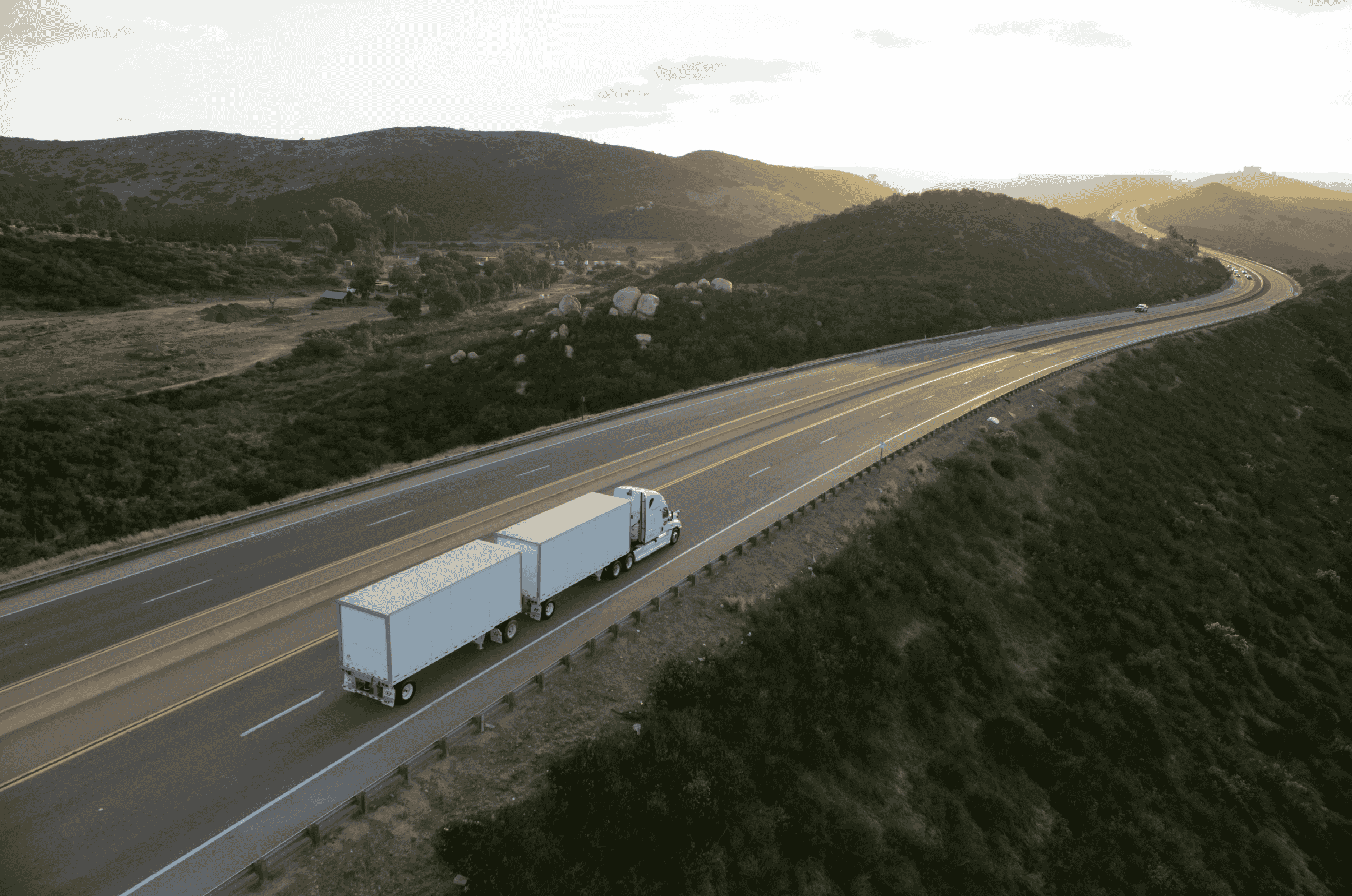 An aerial view of a semi-truck pulling two white Hyundai Translead trailers on a highway through a hilly landscape, featuring the HT TEMPS logo, showcasing Hyundai's innovative trailer technology for thermal event monitoring and heat buildup prevention.